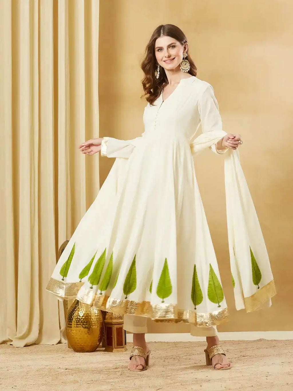 Woman wearing an elegant cream Anarkali suit with green leaf print and gold border.