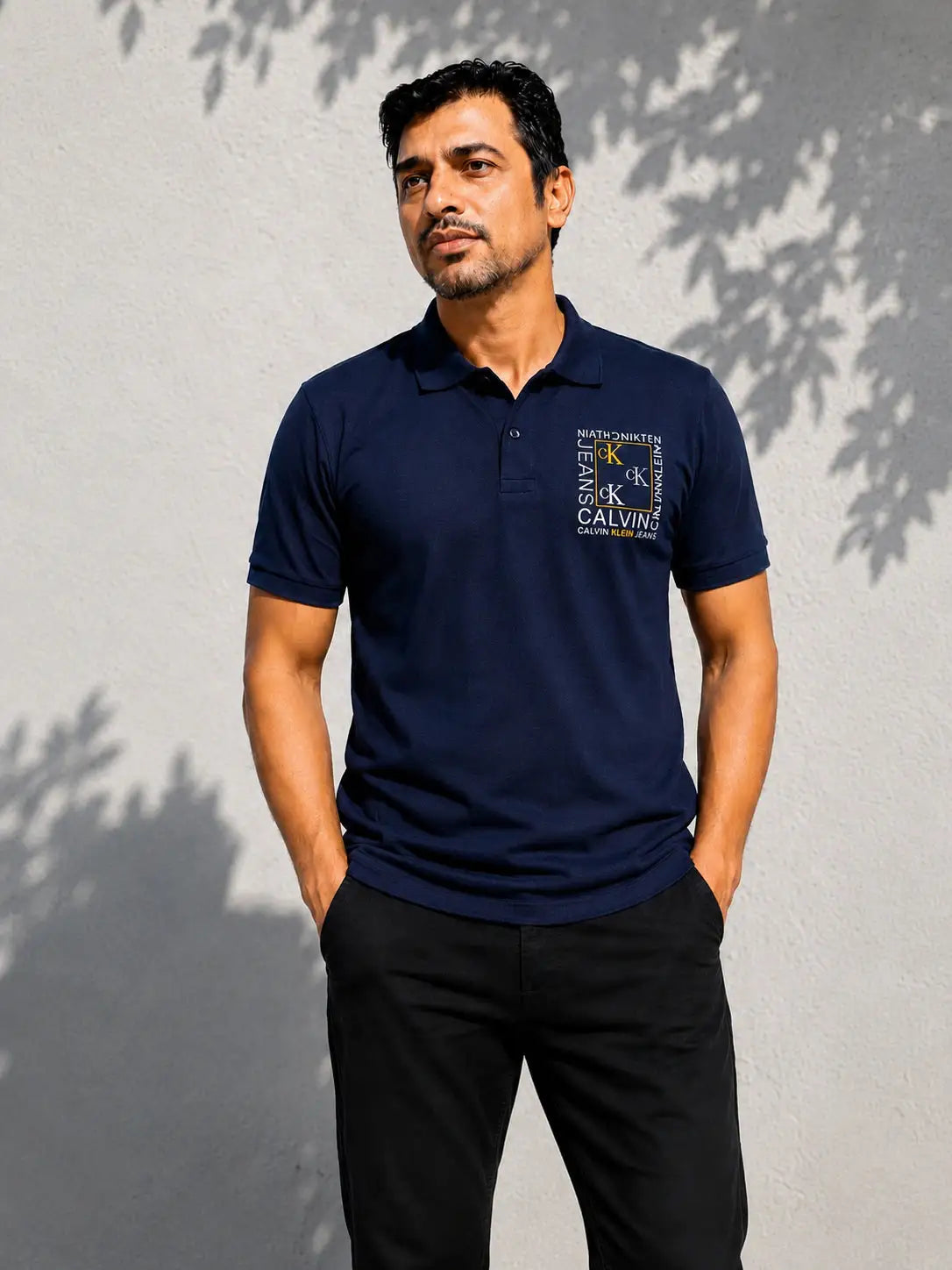 Man wearing a navy blue Calvin Klein polo shirt and black trousers against a grey wall.