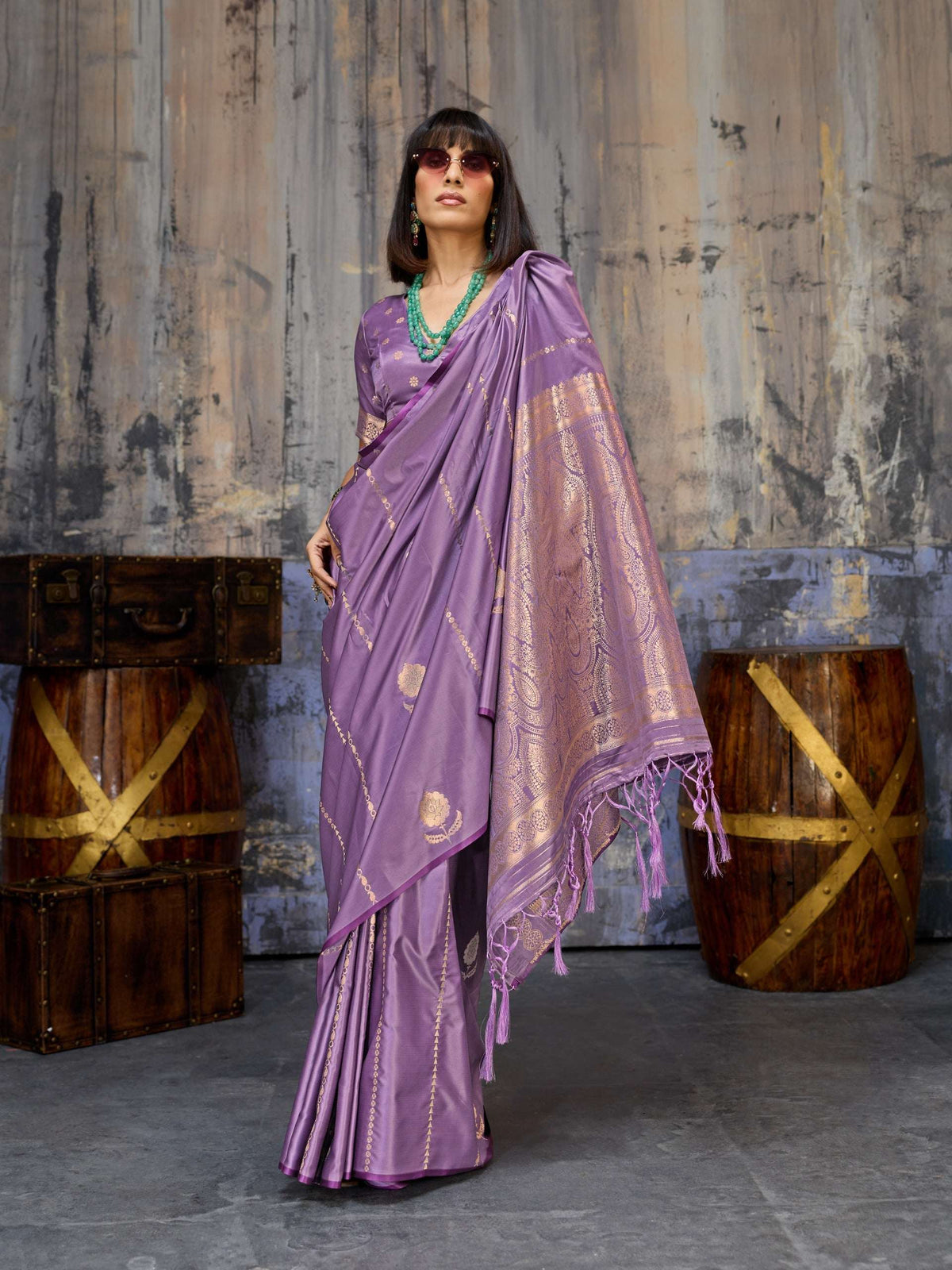 Woman wearing a luxurious purple silk saree with gold zari borders and emerald jewelry.