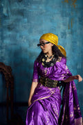 Woman in purple silk saree with gold embroidery, yellow headscarf, and chunky black necklace.