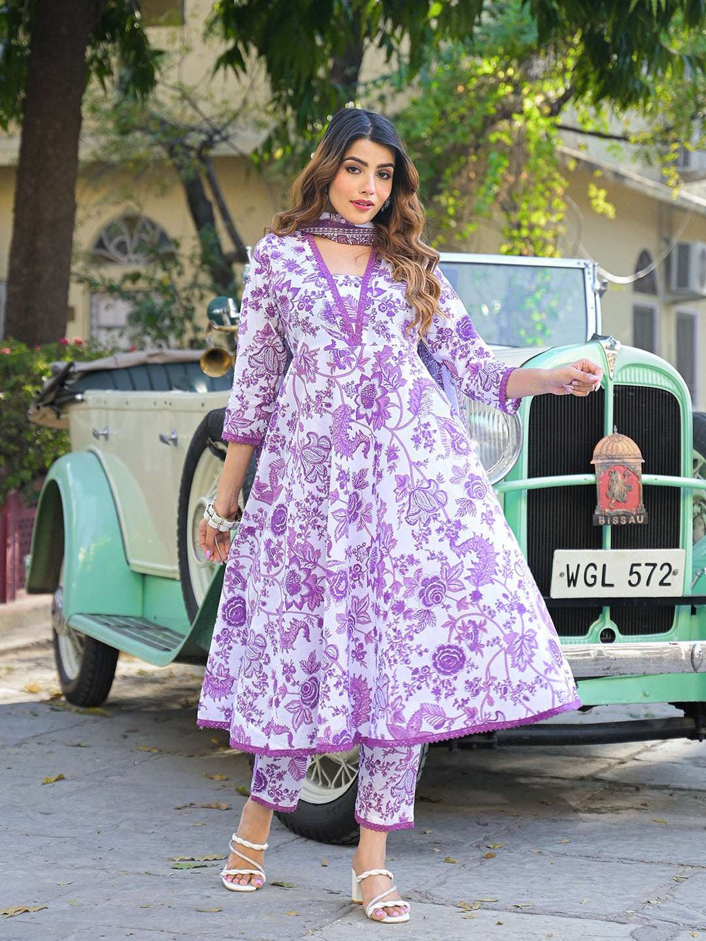 Woman wearing a purple and white floral print Indian ethnic Anarkali suit standing by a vintage car.