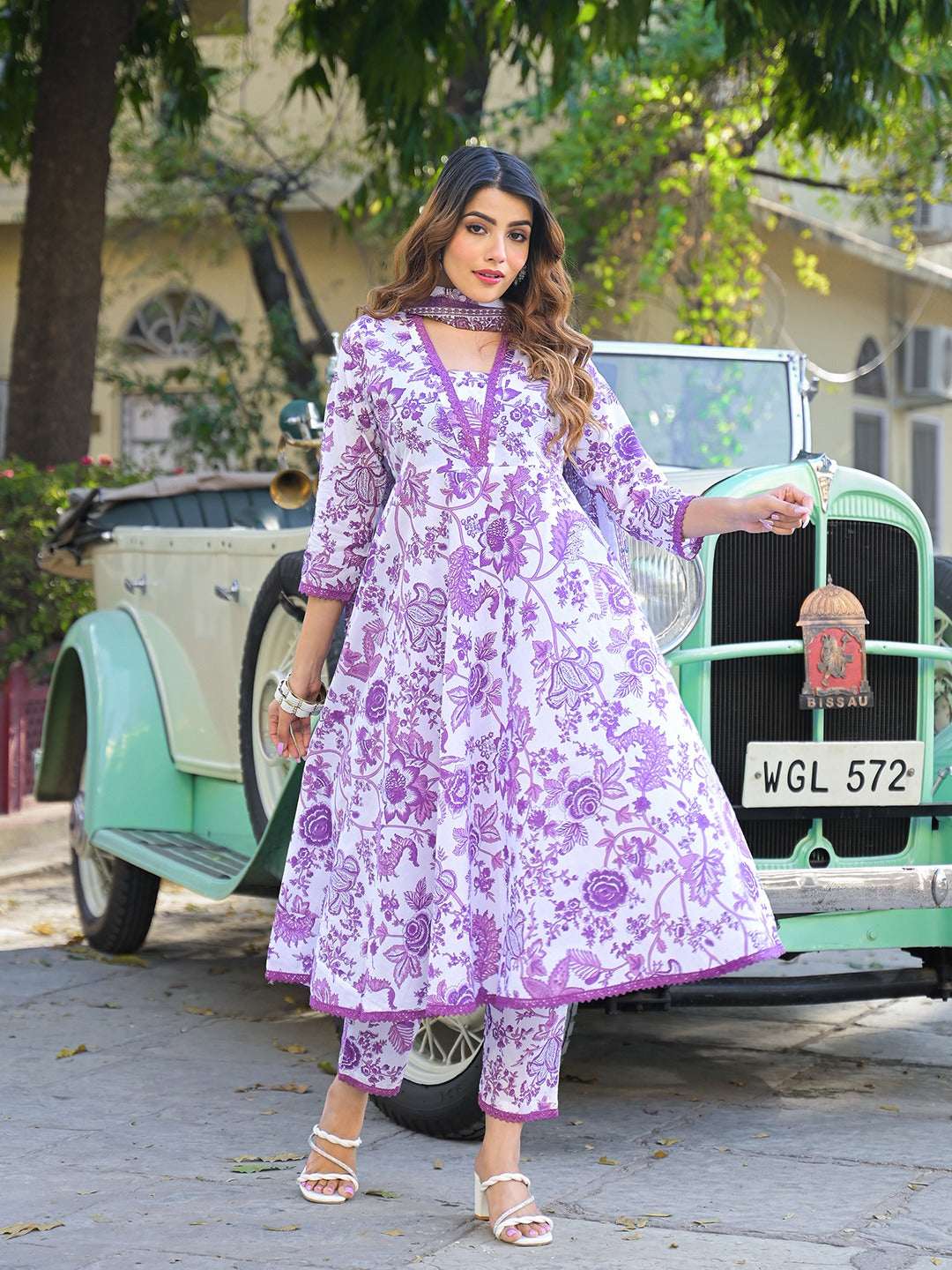 Woman wearing a purple and white floral print Indian ethnic Anarkali suit standing by a vintage car.