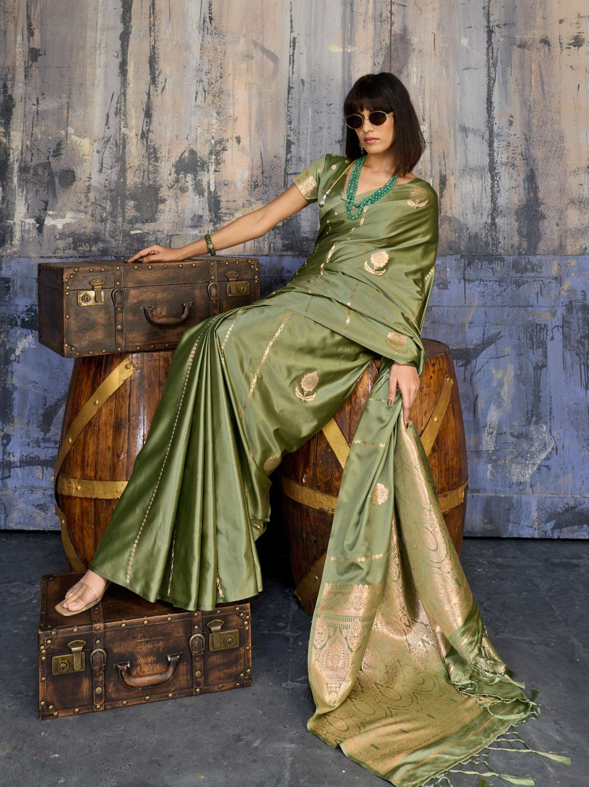 Woman in olive green silk saree with gold zari work sitting on wooden barrels.