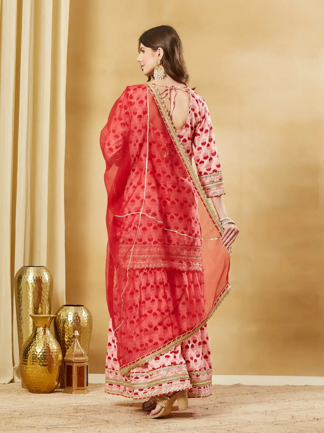 Woman in pink floral print ethnic sharara suit with red dupatta and gold jewelry.