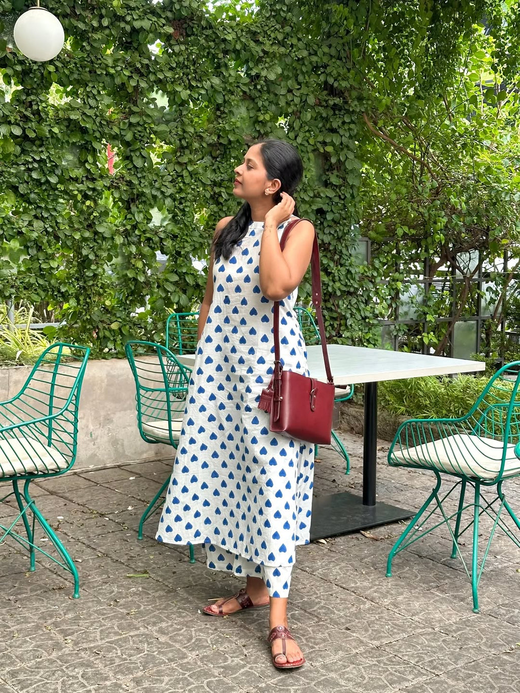 Woman wearing a white and blue printed ethnic dress with a maroon handbag in a garden setting.