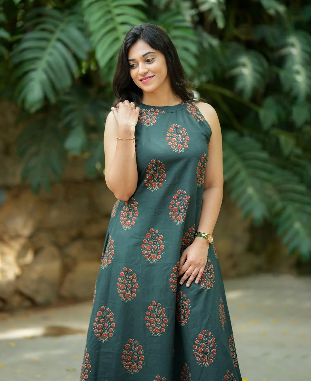 Woman wearing a dark green sleeveless ethnic dress with red floral prints against a leafy backdrop.